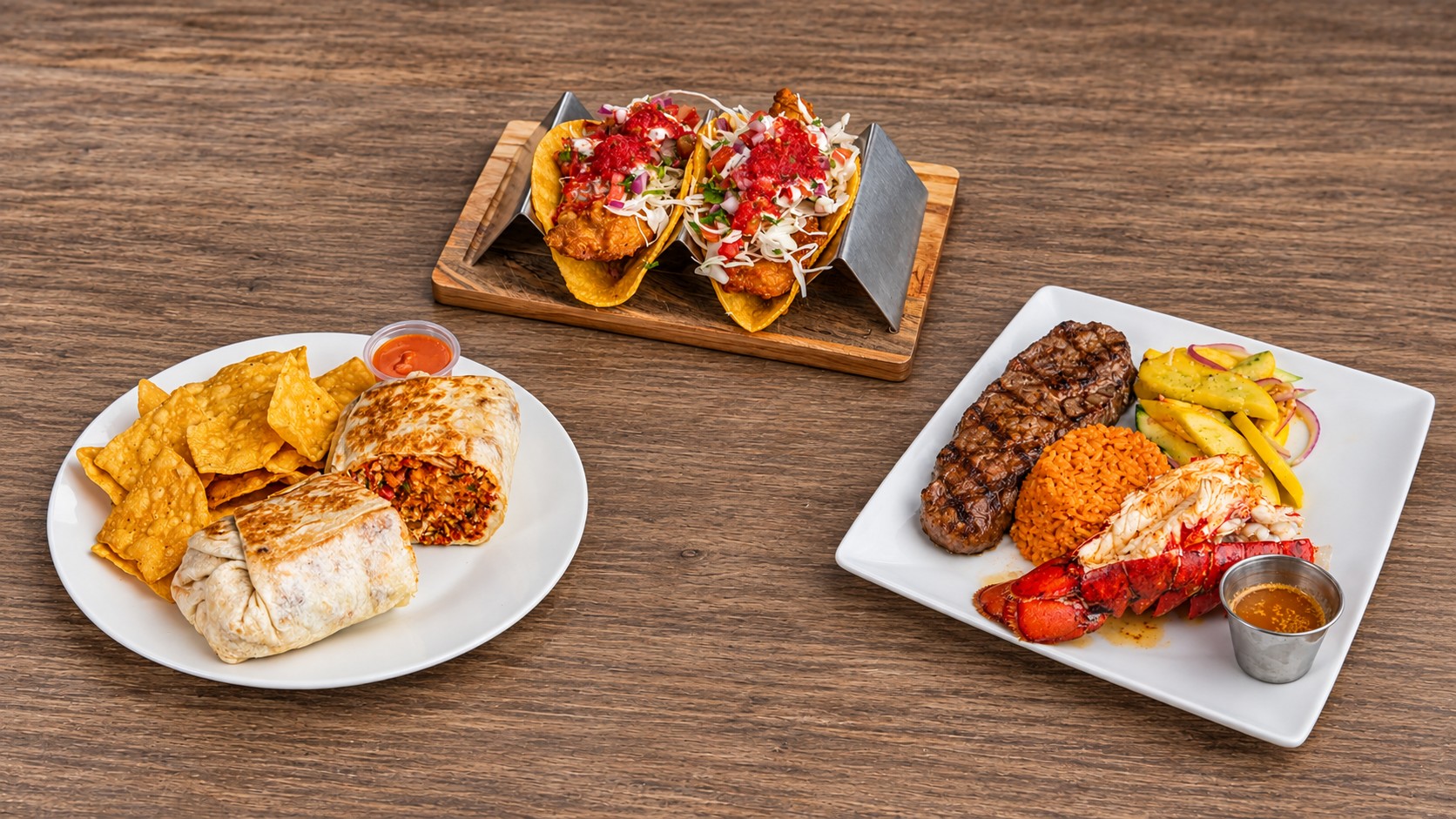 Battered fish tacos in a taco rack, a burrito with chips and salsa, and a steak and lobster tail plate on a wood table