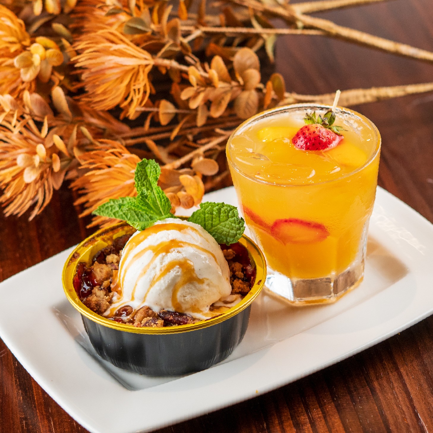 A warm fruit cobbler topped with vanilla ice cream and mint beside an orange cocktail, with dried autumn flowers behind