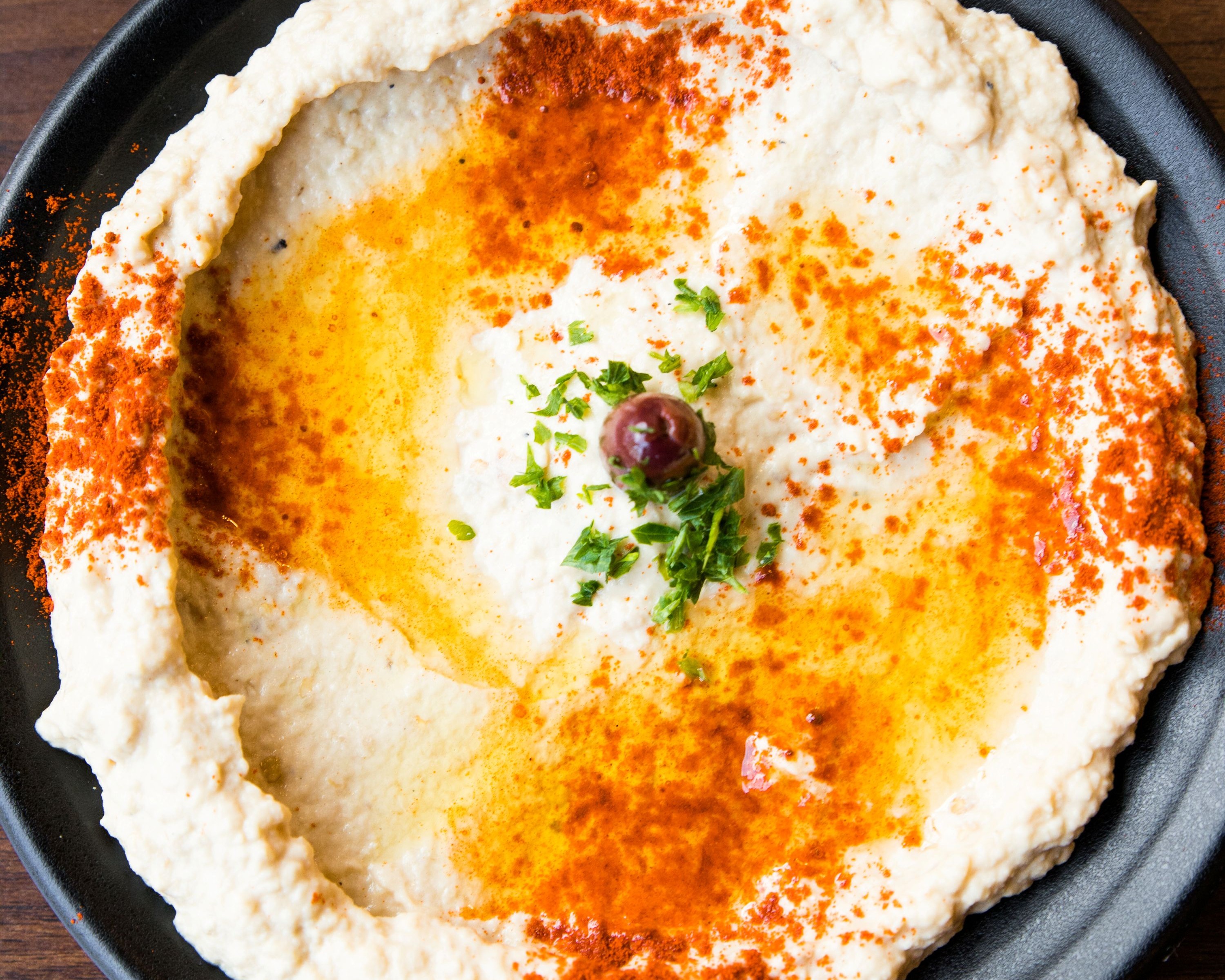 Close-up of hummus in a black bowl with olive oil, paprika, parsley and an olive