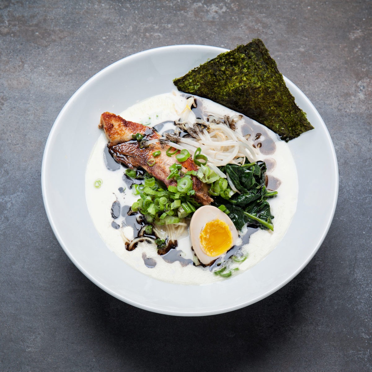 A bowl of The Classic tonkotsu ramen from above: creamy pork broth, chashu, spinach, black garlic oil, a soft egg and nori on slate