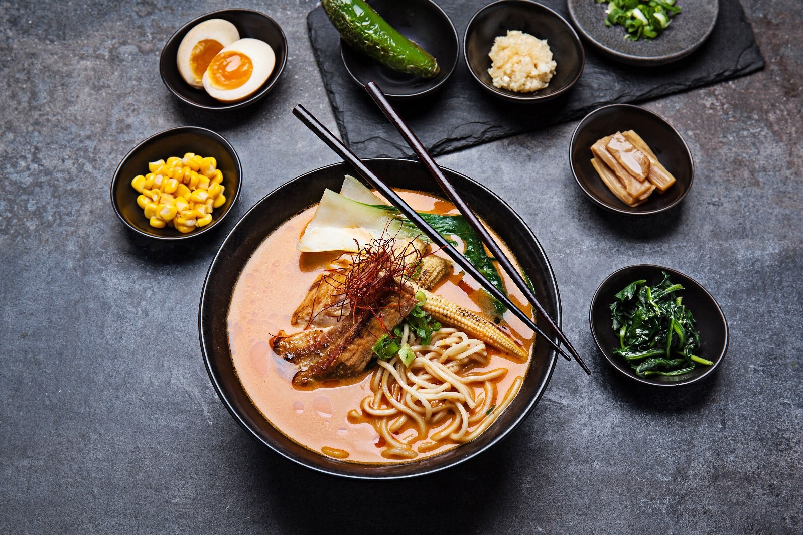 A bowl of spicy ramen with chopsticks resting across it, ringed by small dishes of egg, corn, greens and pickles on a dark slate table